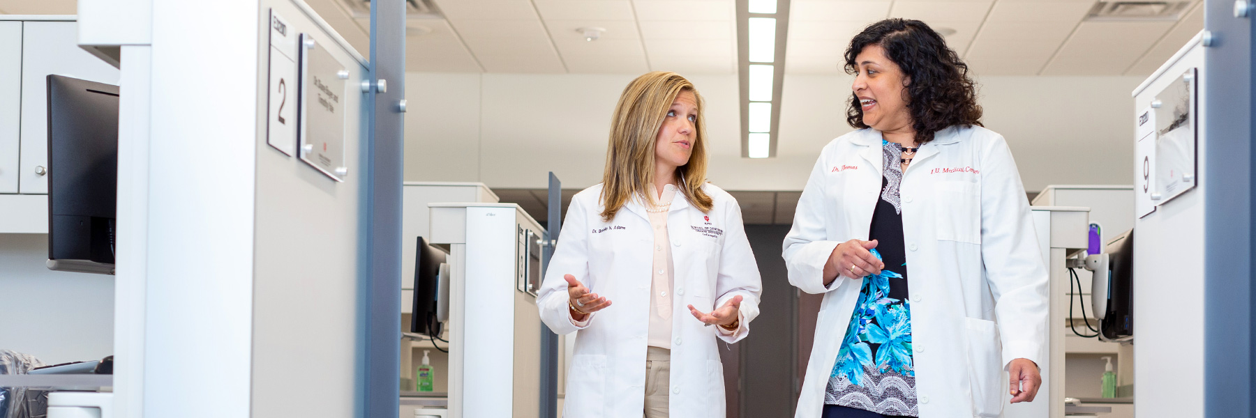 Two female dental professionals in lab coats walk down a hall past exam stations.